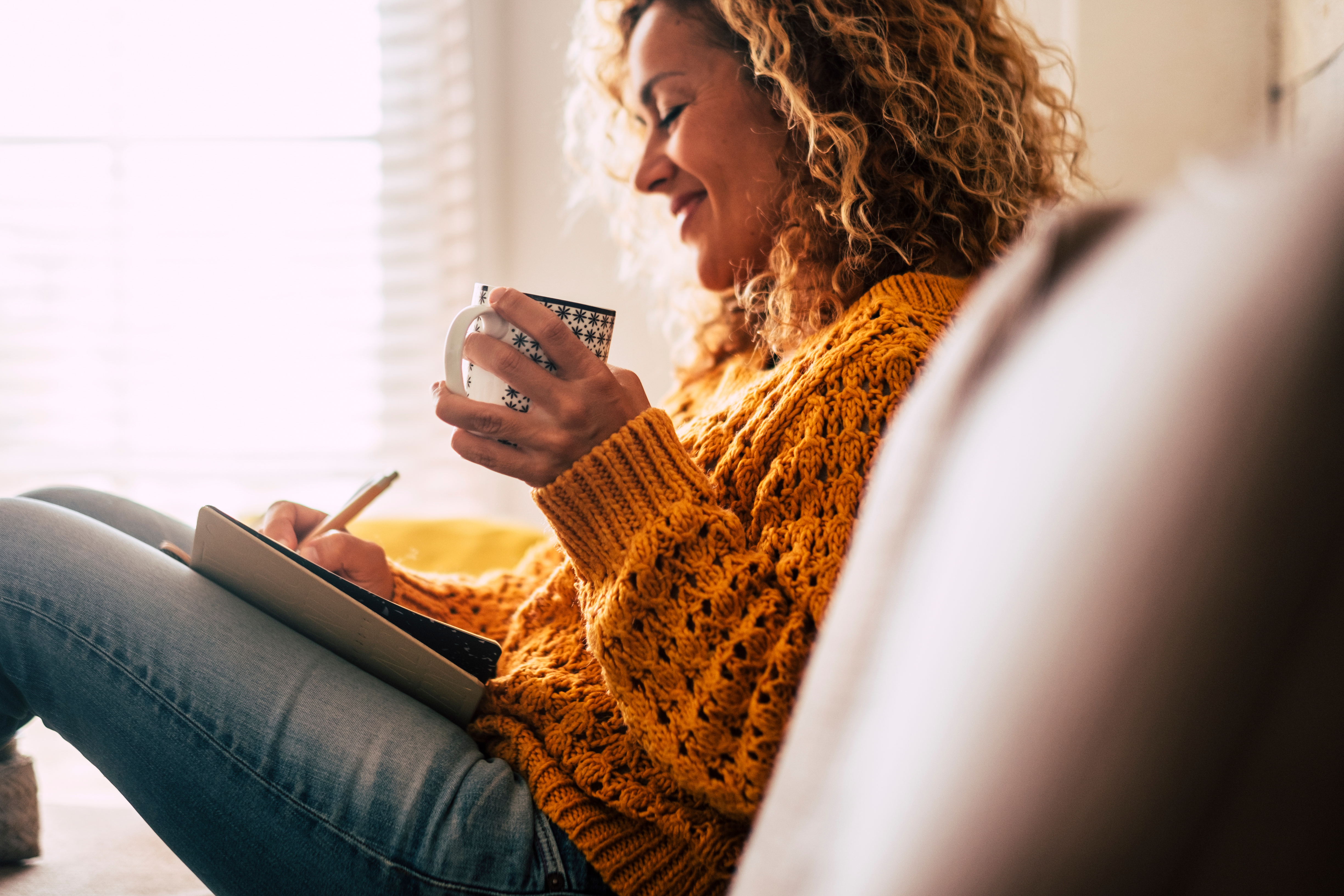 Woman sitting on a couch looking at a tablet and drinking coffee.